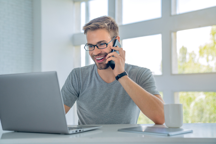 Smiling handsome man in eyeglasses talking by smartphone and using laptop computer while sitting by the table at office